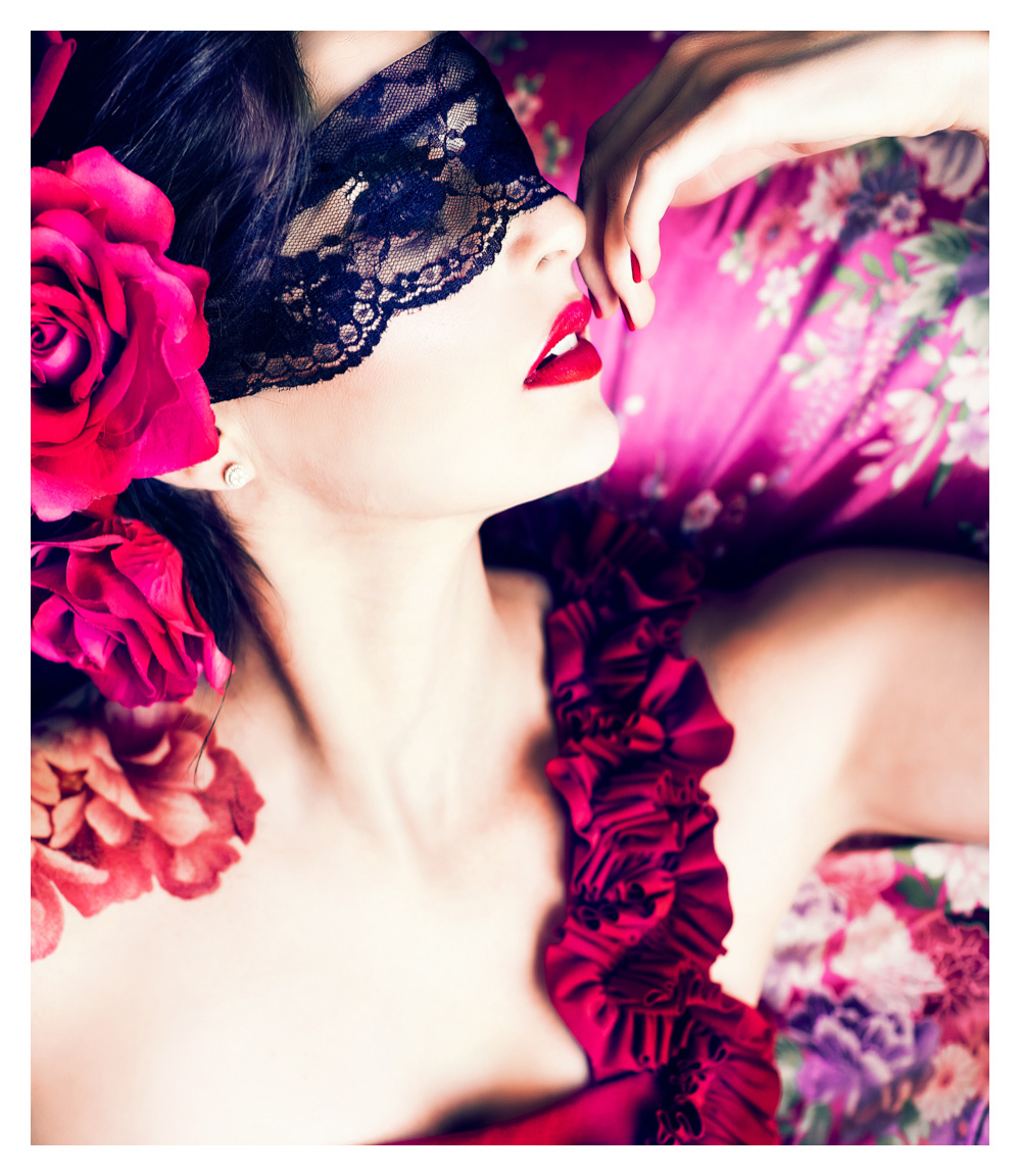 Model in red dress with flowers in her hair poses in black mask during halloween fashion photography shoot