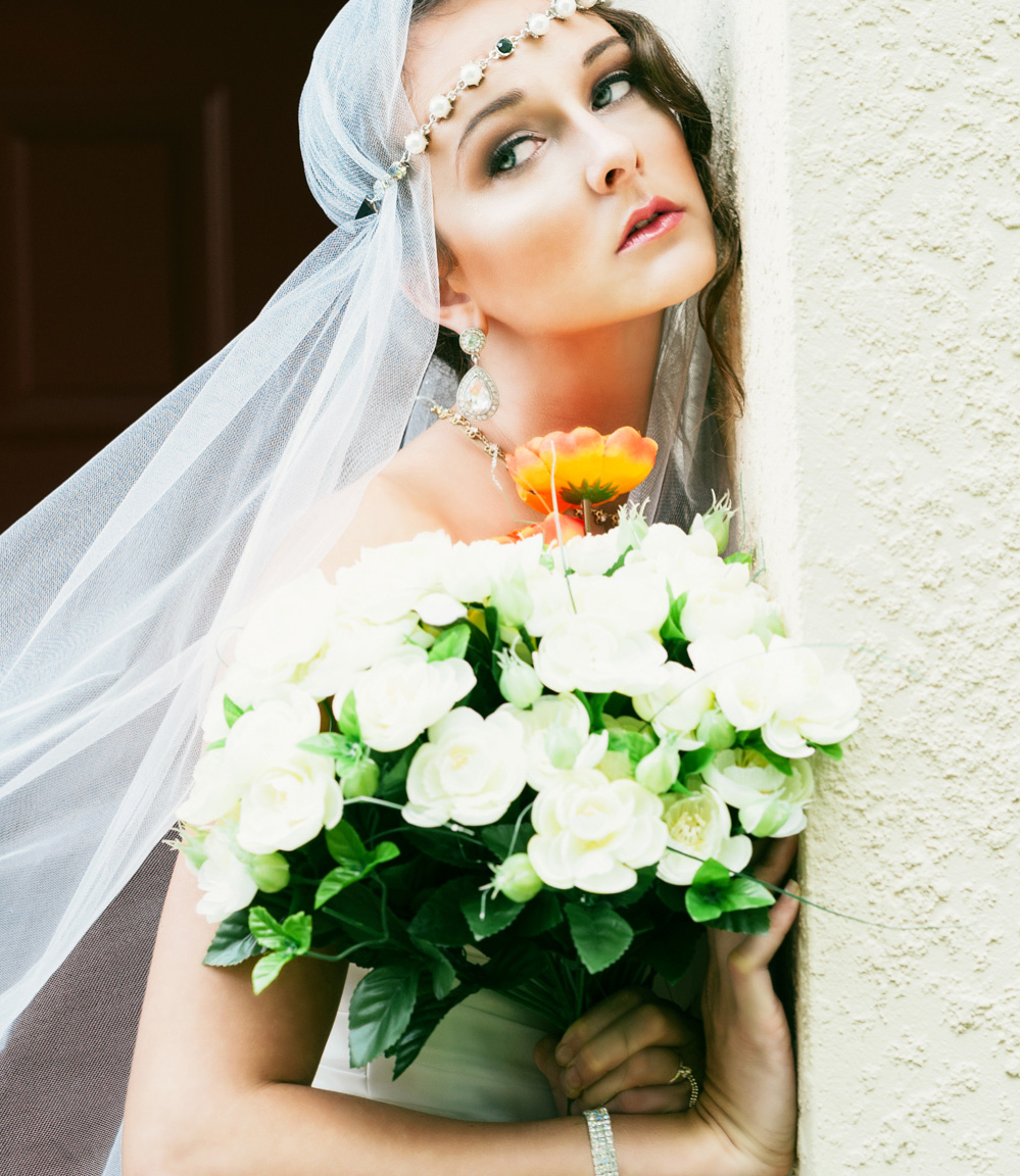 The wind blows the veil of this bride holding flowers near a white wall.