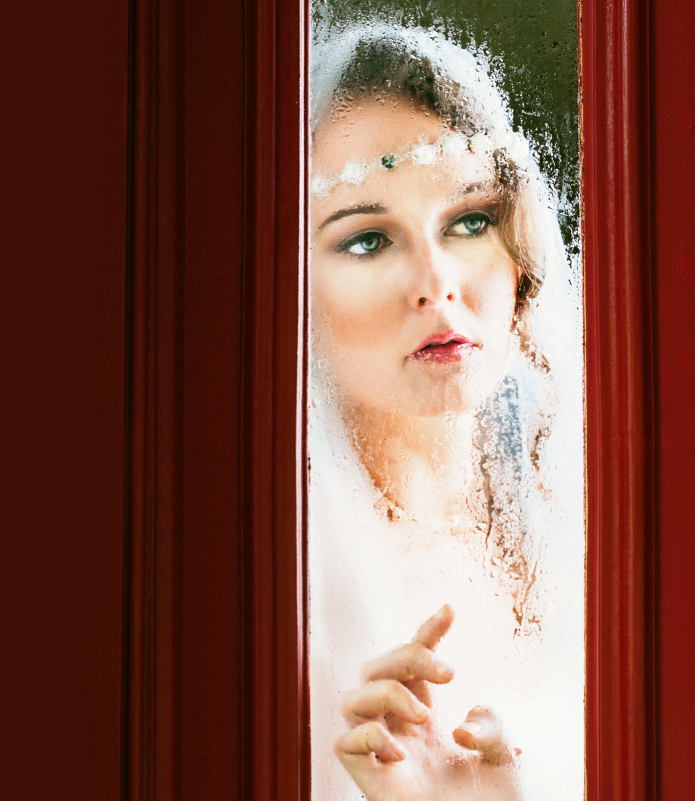 A bride in white looks out a fogged window into the garden